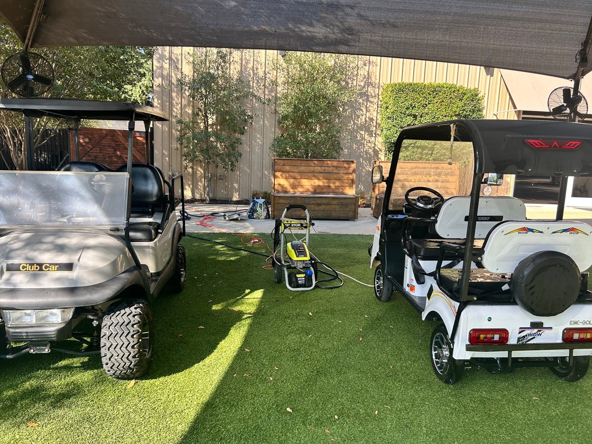 Three golf carts parked in a row on artificial turf under a covered area with metal buildings in the background