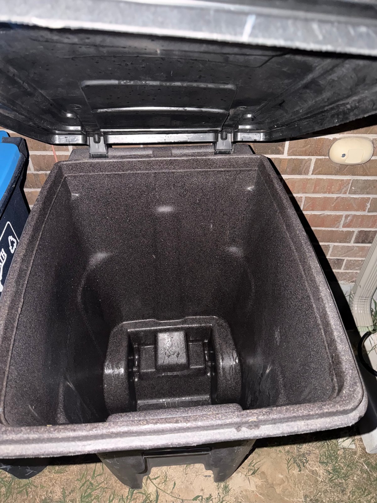 Open dark garbage bin with metal locking mechanism inside, positioned against brick wall