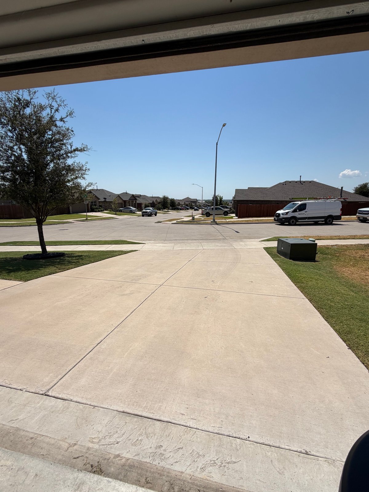 View from under a covered structure of a wide driveway with green grass, parked vehicles, and suburban landscape under clear sky